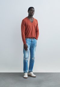 Jeune homme debout contre un mur gris portant un pull orange à col en V, un jean bleu clair déchiré, une ceinture marron et des baskets blanches.