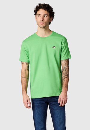 Man met krullend haar, die een felgroen T-shirt met korte mouwen en een klein planetenlogo op de borst draagt, en blauwe spijkerbroek, staande voor een effen achtergrond.