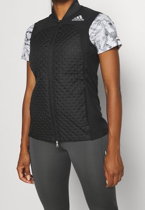 Kvinde iført sort quiltet Adidas ærmeløs vest over hvid mønstret kortærmet skjorte og mørkegrå leggings, stående foran ensfarvet baggrund.