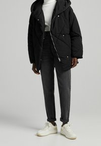 Czarna pikowana kurtka puchowa z kapturem i srebrnym zamkiem błyskawicznym, noszona na białym prążkowanym topie, zestawiona z ciemnymi dżinsami i białymi trampkami.