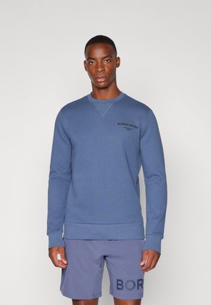 Blå sweatshirt med ribbede ærmer og kant, med et lille sort logo på brystet. Båret med lysegrå shorts, der har fed skrift.