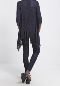 Tmavě námořnický otevřený kardigan s tříčtvrtečními rukávy, zdobený třásněmi na spodním lemu a jemnými horizontálními pruhy, kombinovaný s úzkými džínami a černými botami.