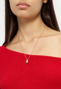 Γυναίκα που φοράει ένα κόκκινο πουλόβερ χωρίς ώμους και ένα λεπτό χρυσό κολιέ με μενταγιόν σε σχήμα δακρύου.
