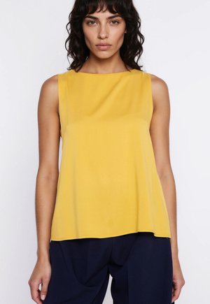 Kobieta o ciemnych falowanych włosach, ubrana w żółty bezrękawnik i ciemnoniebieskie spodnie, na jednolitym białym tle.