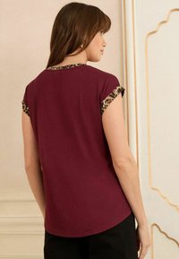T-shirt bordeaux con rifiniture in stampa leopardata sulle maniche. Tessuto morbido, orlo arrotondato e maniche corte. La vista posteriore mostra i dettagli del colletto.