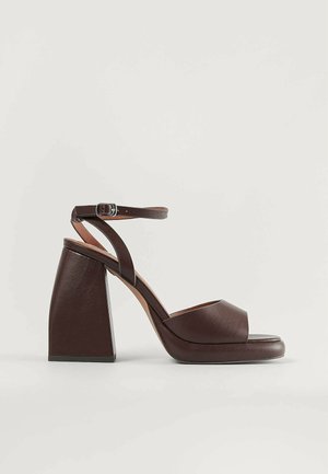 Brown leather heeled sandals feature a wide toe strap, ankle strap with a buckle, and a chunky angular block heel. Smooth texture overall.