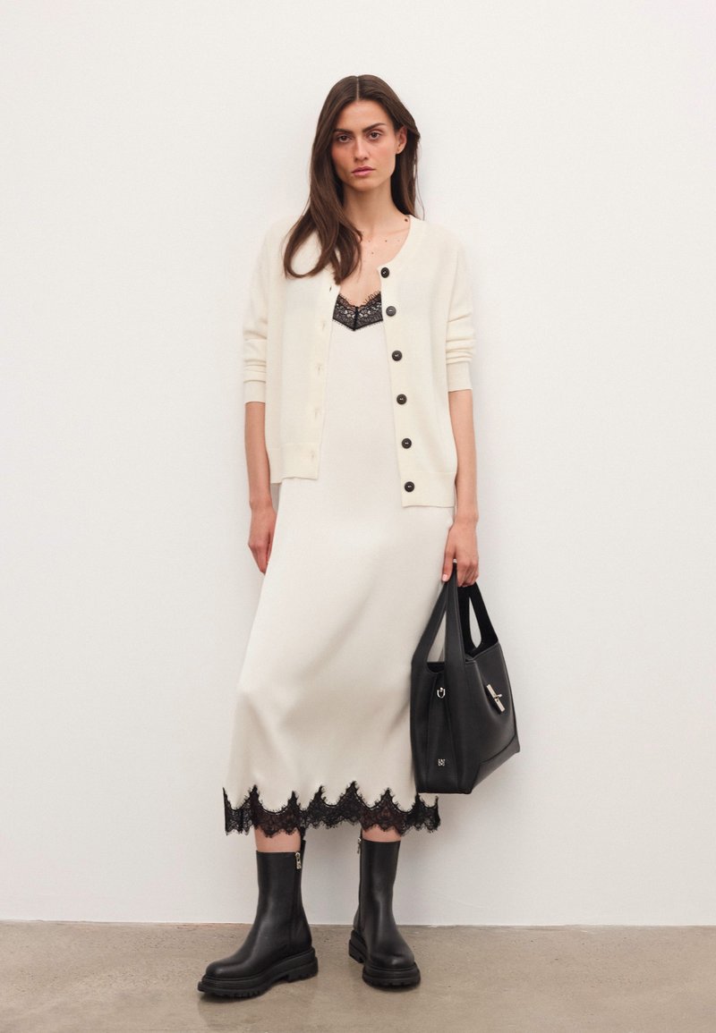 Sieviete, kas ģērbusies krēmkrāsas kleitā ar melnu mežģīņu apdari, krēmkrāsas pogātā džemperī, melnās zābakos un turot melnu rokassomu, stāv pretī baltai siena.