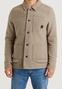 Beige ullblandad jacka med krage, fem knappar, två bröstfickor och två nedre fickor. Har en logotyp-patch på vänster bröst.