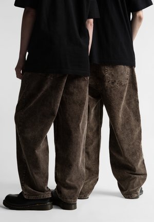 Due persone che indossano pantaloni di velluto a coste marroni larghi, magliette nere e scarpe nere, in piedi fianco a fianco, di spalle su uno sfondo bianco.