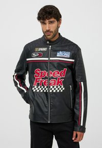 Giacca da corsa in pelle nera con spalle imbottite, strisce rosse e bianche, e grafica "Speed Freak" con accenti a motivo a scacchi.