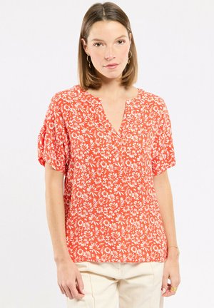 Femme aux cheveux bruns et raides portant un chemisier orange à manches courtes avec un imprimé floral blanc et un pantalon de couleur claire, debout devant un fond uni.