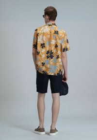 Korte mouw gele bloesemshirt met marineblauwe en lichtblauwe patronen, gecombineerd met marineblauwe shorts. Het model draagt bruine schoenen en houdt een marineblauwe pet vast.