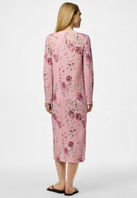 Lång rosa klänning med blommönster, med veckad textur, långa ärmar och nyckelhål i ryggen. Bärs med platta sandaler.
