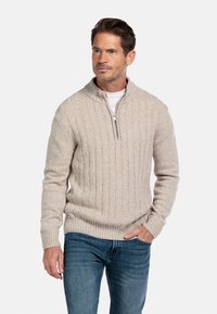 Lichtbeige gebreide trui met een halflange ritssluiting, voorzien van een geribbelde textuur en aansluitende manchetten, gecombineerd met blauwe jeans.