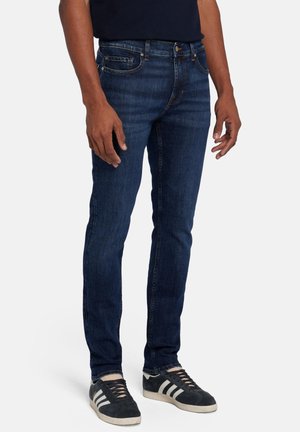 Jeans in denim blu scuro con vestibilità affusolata, design a cinque tasche e dettagli di cucitura sottili, abbinati a scarpe nere con strisce bianche.