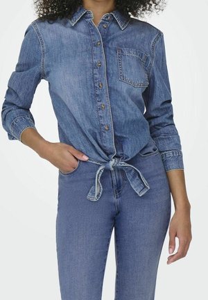 Frau trägt ein geknöpftes blaues Jeanshemd, das an der Taille gebunden ist, und passende blaue Jeans, steht mit einer Hand in der Tasche.