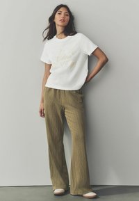 T-shirt en coton blanc avec le texte brodé "Le Dolce Vita", associé à un pantalon large à motifs verts. Le mannequin porte des chaussures de couleur claire.