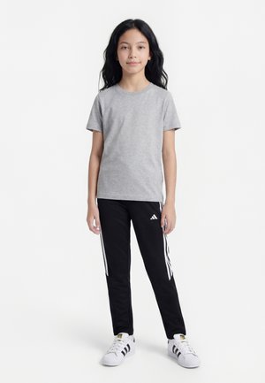 Bambino con lunghi capelli neri che indossa una maglietta grigia a maniche corte, pantaloni da tuta neri Adidas con strisce bianche e scarpe da ginnastica bianche Adidas.