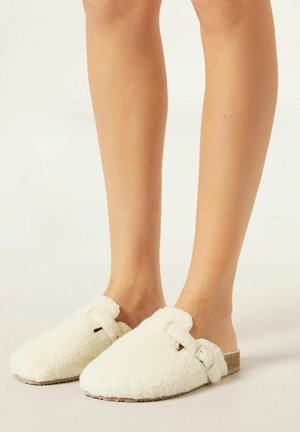 Chaussons blancs en peluche avec une texture douce, design slip-on et une seule sangle avec une petite boucle. La semelle est en matériau naturel.