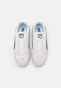 Weiße Sneaker aus Wildleder und Leder mit schwarzen Akzenten, flachen Schnürsenkeln und einer hellblauen Innensohle. Puma-Logo auf der Zunge und Seite.