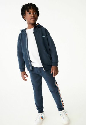 Tumesinise lukuga kapuutsiga särk, millel on ees taskud ja tikandiga logo, koos sobivate pükstega, mille külgedel on mitmevärvilised triibud. Valged tossud.