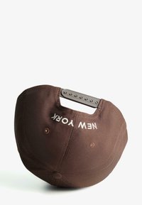 Brown baseball cap with a rounded crown, featuring a white "NEW YORK" logo and a plastic strap closure at the back.