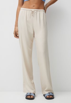 Femme portant un pantalon beige à larges jambes avec cordon et des sandales bleues, debout devant un fond clair uni.