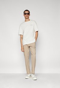 Witte katoenen t-shirt met korte mouwen en tekstdetails, gecombineerd met beige cargobroek en witte sneakers. Model draagt een zonnebril.