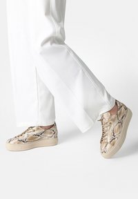 Persona con pantalones blancos y zapatillas con cordones beige con estampado de serpiente sobre un fondo blanco liso.