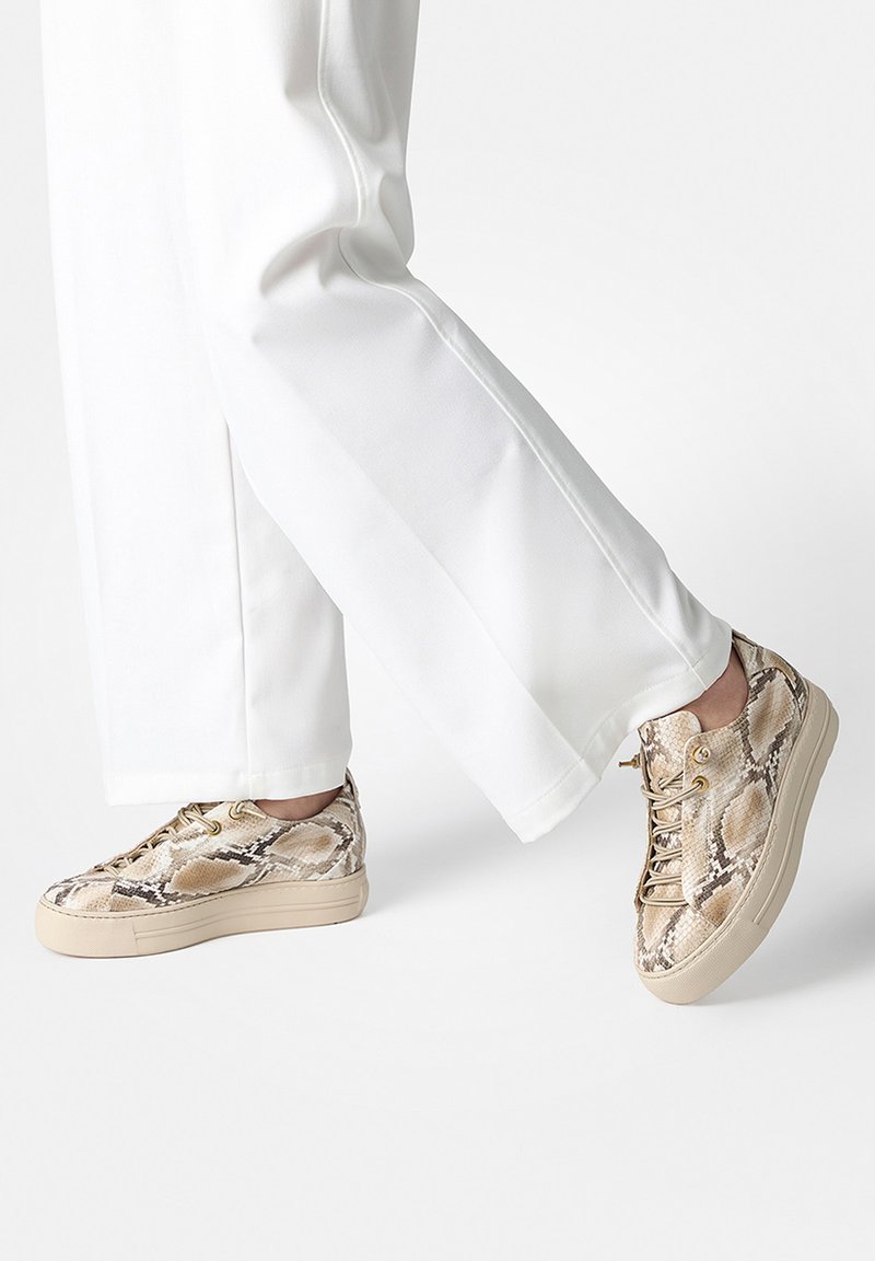 Persona con pantalones blancos y zapatillas con cordones beige con estampado de serpiente sobre un fondo blanco liso.