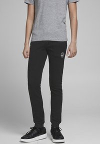 Schwarze, taillierte Jogginghosen mit elastischem Bund, weichem Stoff, seitlichen Taschen und einem runden Logo auf dem linken Oberschenkel, kombiniert mit einem grauen T-Shirt und schwarzen Sneakern.