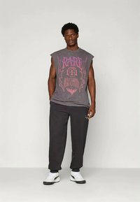 Vīriešu modelis ģērbies melnās biksēs, bez piedurknēm tumši pelēkā grafiskā T-kreklā un baltās sporta apavos, stāv pretī vienkāršai gaišai fona.