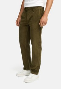 Pantalones de pana verde oliva con diseño de pierna recta, cuentan con pliegues delanteros y un pequeño logo en el costado, combinados con zapatillas blancas.