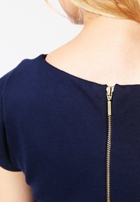 Námorně modrý top s hladkou texturou, kulatým výstřihem a zlatým zipem na zádech, který přidává funkční detail.