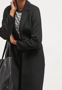 Černý dlouhý kabát se strukturovaným designem, vyrobený z hladkého materiálu. Má šálový límec a zapínání vpředu, doplněný černou kabelkou.