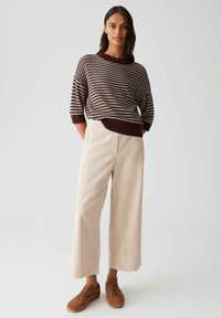 Woman standing with hands in pockets wearing striped brown sweater, beige wide-leg pants, and brown sneakers on plain light background.