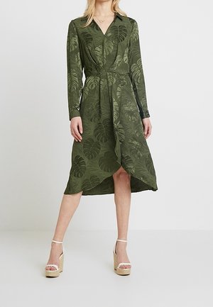 Kobieta ubrana w oliwkową, długorękawową sukienkę kopertową z wzorem liści oraz beżowe sandały na platformie, stojąca na jednolitym jasnoszarym tle.