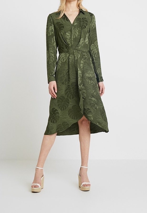 Femme portant une robe portefeuille à manches longues vert olive avec un motif de feuilles et des sandales compensées beiges, debout devant un fond gris clair uni.