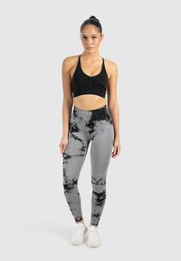 Top sportivo nero con dettagli arricciati, abbinato a leggings grigi con un motivo marmorizzato nero. Modella in piedi con scarpe da ginnastica bianche.