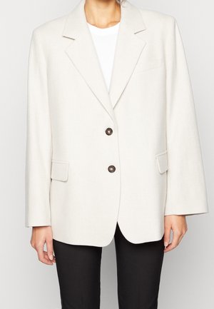 Blazer beige en tissu texturé avec un col cranté, deux boutons et poches latérales, associé à un haut blanc et un pantalon noir.