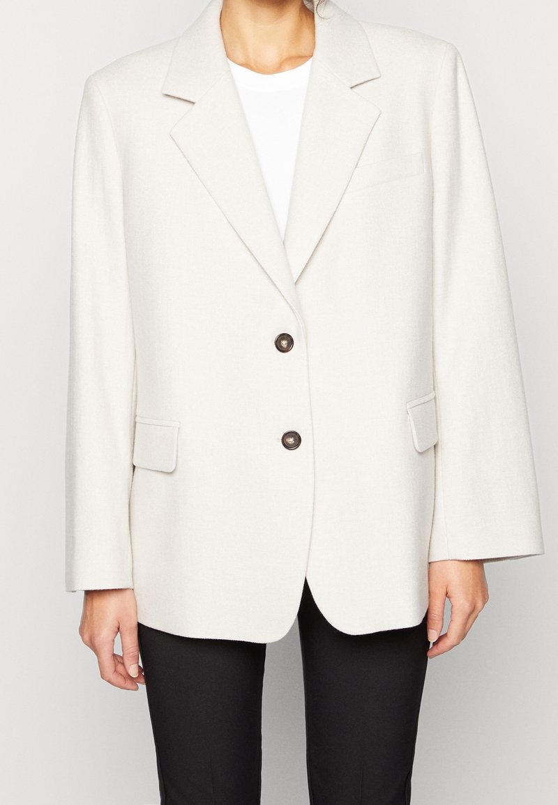 Blazer beige realizzato in tessuto testurizzato con rever a forma di punta, due bottoni e tasche laterali, abbinato a una maglietta bianca e pantaloni neri.