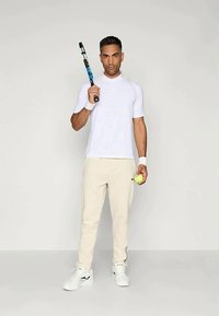 Fehér sportpóló, bézs melegítőnadrág, teniszkészletet és labdát tart a kezében. A fehér sportcipők teszik teljessé az öltözéket. Egyszerű dizájn, sima textúra.