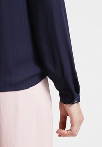 Camicia a maniche lunghe blu navy con orlo arrotondato, dotata di dettaglio a bottoni al polsino, realizzata in tessuto liscio e leggero.