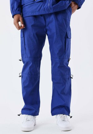 Pantalones cargo azules con múltiples bolsillos, puños ajustables y una tela ligera. Combinados con zapatillas blancas para un aspecto limpio.