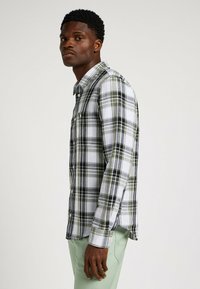 Koszula w kratę w kolorze białym, szarym i oliwkowym, z kołnierzykiem zapinanym na guziki i długimi rękawami. Zauważalny detal kieszeni po lewej stronie.