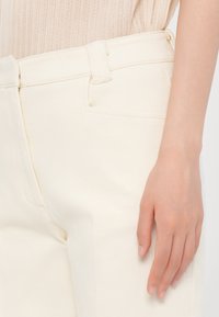 Gros plan d'une personne portant un pantalon écru avec une boucle de ceinture visible et une main reposant à côté de la cuisse.