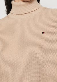 Beżowy sweter z żebrowanym splotem i golfem, z małym prostokątnym logo w kolorach czerwonym, białym i granatowym na klatce piersiowej, noszony przez osobę z ciemnymi włosami.