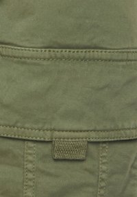 Olivgrön tyg med en slät textur, med en ficka och sydda sömmar. En vävd etikett syns nära fickområdet.