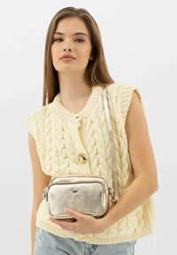 Metalická zlatá crossbody taška s texturovaným povrchem. Obdélníkový tvar s uzavíráním na zip a odnímatelným popruhem. Nosená ležérně proti krémovému svetru.