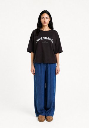 Frau, die ein schwarzes übergroßes T-Shirt mit dem Text "Copenhagen", weite blaue Jeanshosen und braune Schuhe vor einem weißen Hintergrund trägt.